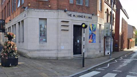 Street view image of the Making Rooms building in Blackburn. The ground floor is white brick and the top floor is red brick. It has long windows and a bright poster next to the black front door and stands next to a pedestrian crossing.