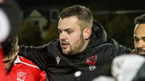 Elliot Powell flanked by players either side is talking in a huddle. He is wearing a black Jersey Bulls coat with the initials EP on.
