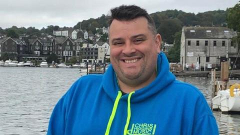 Chris Higgins stands in front of a harbour and is wearing a bright blue hoodie with green strings and a green logo bearing his name.