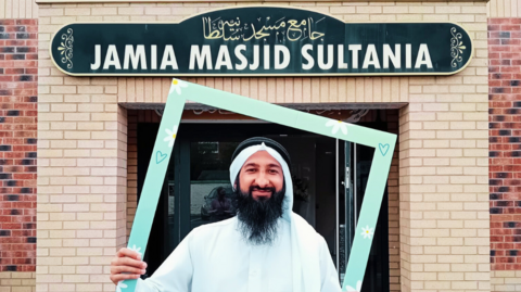 Imam Habeeb Minhas in front of the mosque which is built from red and sand-coloured brick. The imam has a long black beard and is wearing traditional clothing