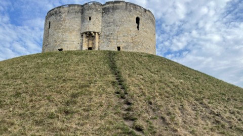 A curved stone tower on a dry grassy mound. Blue skies with clouds in the background. The mound has visible marks of where people have walked up and down.