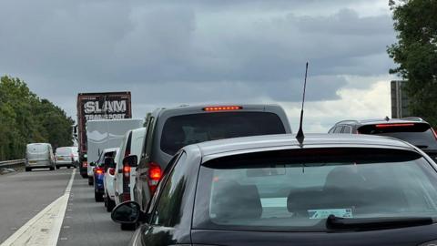 The dual carriageway of the A1 where a slip road is merging onto the carriageway. There are cars and lorries queuing on the road.