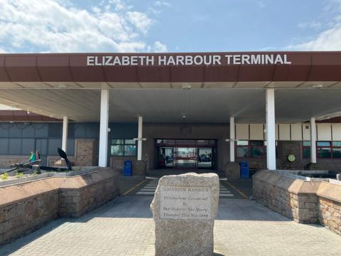 Entrance to Elizabeth Harbour Terminal on a sunny day. An engraved rock says 'Elizabeth Harbour was opened by Her Majesty The Queen Thursday 25 May 1989'. Large glass double doors are open into the terminal.