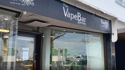 The picture shows the front of The VapeBar in Guernsey. The shop sits on the ground floor of a white building, and the black sign above the entrance reads “The VapeBar guernsey” in white lettering. 