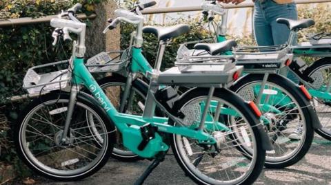 Three Beryl Bikes parked in a bay. They are turquoise in colour, with baskets at the front. They are next to a barrier, with the sea just glimpsed beyond. A woman is stood by the bikes, her top half out of shot. She's wearing jeans and Converse-style trainers.