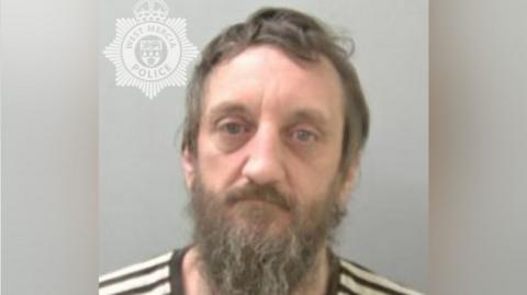 A police mugshot of a man with brown hair and a long brown beard with grey streaks. He is wearing a black and white striped top.