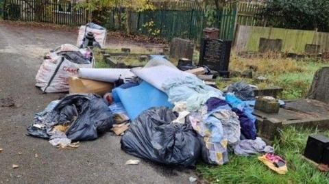 A pile of rubbish is strewn over several graves that have dark or black-coloured headstones. The piles include black bin bags, clothing, cardboard boxes and furniture.