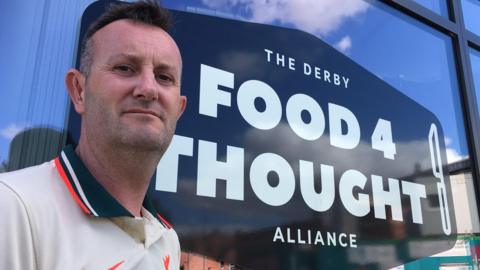 Paul Brookhouse looking at the camera in front of a Derby Food 4 Thought Alliance sign. He has short dark hair, greying at the temples and a light dusting of dark stubble. He 