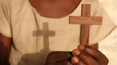 A close up of someone holding a small wooden crucifix with the shadow of the cross on the person's cream T-shirt.