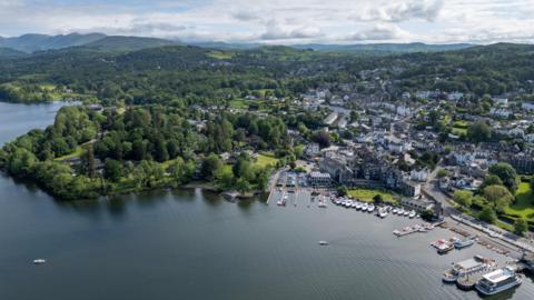 An aerial view of Bowness-on-Windermere, a lake shore town in the Lake District National Park. There are lots of boats moored in the marina and the town stretches out beyond it, surrounded by trees. In the distance are hills and mountains."