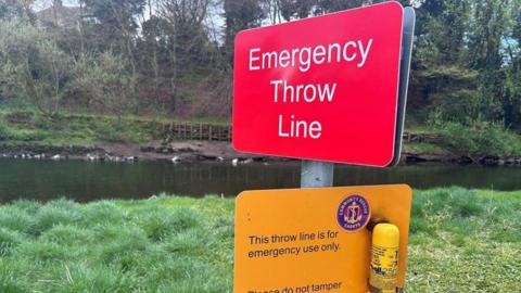 A red emergency throw line sign with a thrown line.
The two signs are red and yellow and placed near an embankment close to a waterway