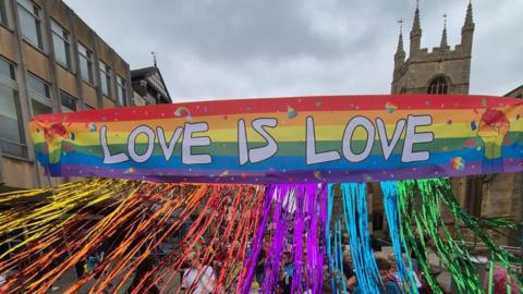 A rainbow-coloured banner has the words "love is love" printed on it and there are streamers coming from the bottom of the banner. A tower can be seen in the background.