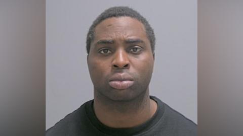 A police custody photo of a man with short dark hair and a black T-shirt.