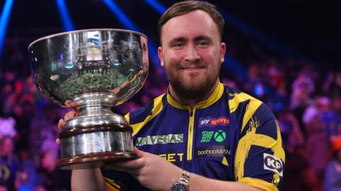 Luke Littler is wearing a yellow and purple top. He faces the camera and lifts the trophy after winning the Grand Slam of Darts. The crowd can be seen behind him. 