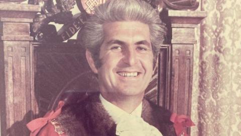 A former mayor wearing his red and black mayoral robes, and the chain of office. 