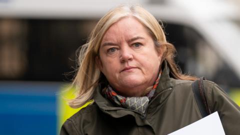 Baroness Casey walks along, wearing a green jacket and grey, red and white scarf. She has shoulder length blonde hair.