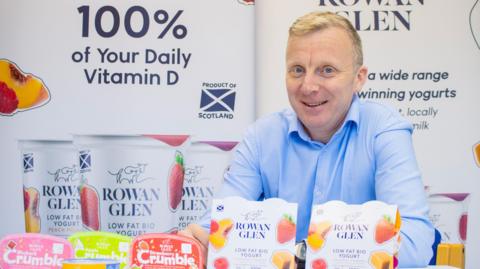 A man in a blue shirt with short blond hair sits behind yoghurt products with the Rowan Glen name on them
