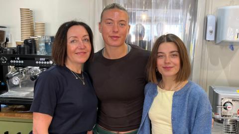 Catherine Bone, Abbie Paul and Izzy Bone stood behind the counter of a bakery, smiling at the camera. Coffee machines are behind them. Catherine Bone has a short brown bob and is wearing a navy T-shirt. Abbie Paul has blonde hair slicked back and is wearing a brown T-shirt. Izzy Bone has a short, brown bob and is wearing a yellow T-shirt and a blue button-up cardigan.