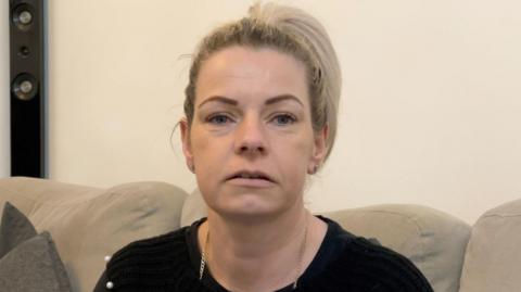 Kerry is sitting on a beige coloured sofa. She has dyed blonde hair, is wearing a black top and is wearing a gold necklace.