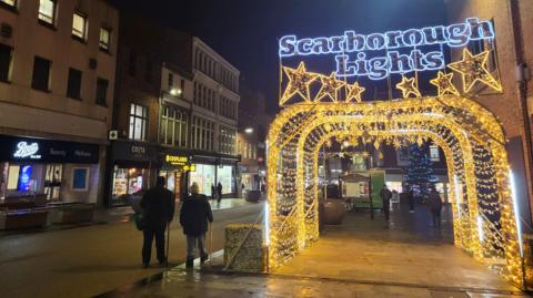A festive street scene at night in Scarborough, featuring a glowing archway that reads “Scarborough Lights” in blue and yellow lights with star decorations.