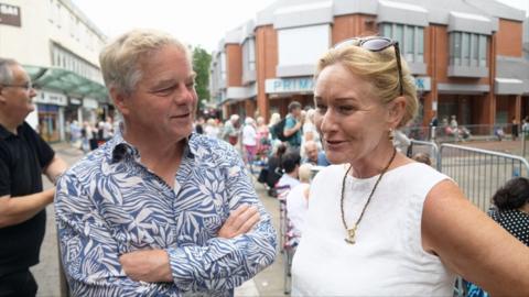 A man and a woman talking. The man has grey hair and a blue and white patterned shirt. The woman has blonde hair, sunglasses on her head and is wearing a white top