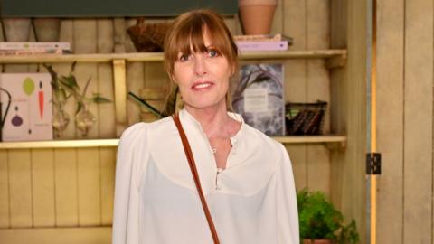 A woman wearing a cream blouse with shelves in the background filled with pots, plants and books
