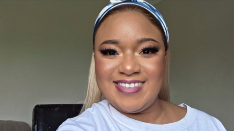 A close-up of Lyn, who is smiling broadly to camera. She has deep pink lipstick and fake eyelashes on. She wears a white T-shirt and has a blue and white bandana on her head. Her straight, blonde hair can be seen falling behind her shoulders.