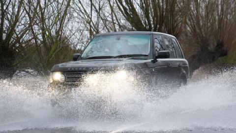 A 4x4 vehicle drives through flood water. Water is spraying into the air as the car drives through it. Trees are in the background behind the car.