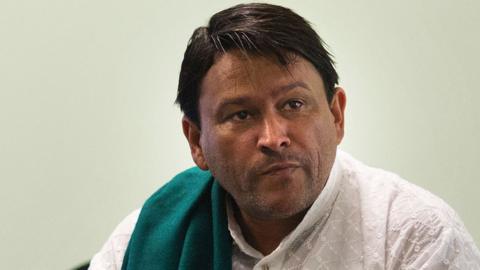 A close-up of Imam Muhsin Hendricks wearing a white shirt and a green scarf draped over one shoulder. The shot was taken in 2016.