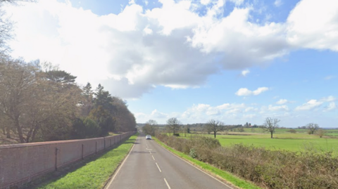 A view of Ridgmont Road. There is a red brick wall running along the left hand side while there is a hedge and fields to the right. A lone vehicle can be seen on the right hand side of the road heading toward the camera.