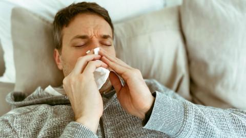A man sitting back on a sofa and blowing his nose with a tissue.