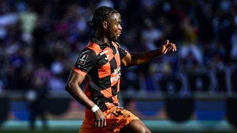 Darlin Yongwa, wearing a black and orange check jersey and orange shorts, dribbles with the ball while playing for Lorient
