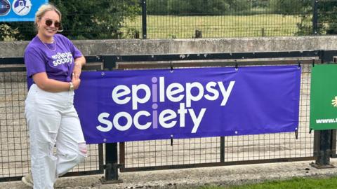 Ellie Thornton smiling for the camera in front of a banner reading Epilepsy Society. She is wearing sunglasses. It is overcast.
