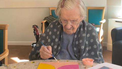 An elderly woman sitting in a wheelchair, wearing a grey spotted shawl over her shoulders, paints a colourful large tile. She is painting blocks of peach, mustard and cerise pink. 