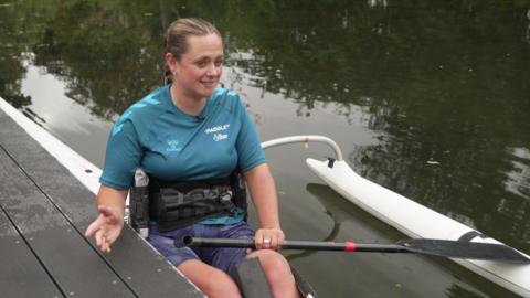A woman with blonde hair sat at a riverside in a white kayak. She's wearing a turquoise short-sleeved top and purple shorts