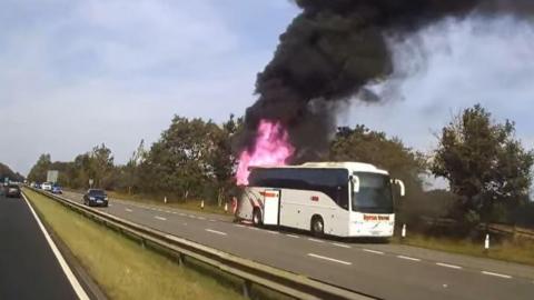 Dashcam footage taken from the other side of the dual carriageway. The white coach has pulled over in the layby and red flames have engulfed the rear section of the vehicle. Large plumes of thick black smoke are billowing in the area. 