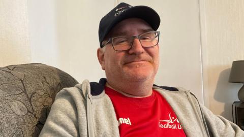 Gleen Faeroy sitting in an armchair, wearing a black baseball cap, red T-shirt and grey track top