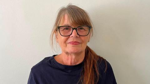 A woman is wearing a dark blue sweater and black glasses looking directly at the camera. her hair is long and falls on her left shoulder. The background is white.