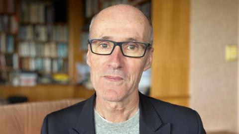 A close up of Jonathan Renouf. He has no hair and is wearing glasses, a dark jacket, and a grey top.
