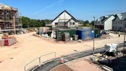 A building site with a house in the middle surrounded by scaffolding. A building on the left is also scaffolded. There is a dust road surrounding the buildings, and a tarmac path at the bottom of the image.