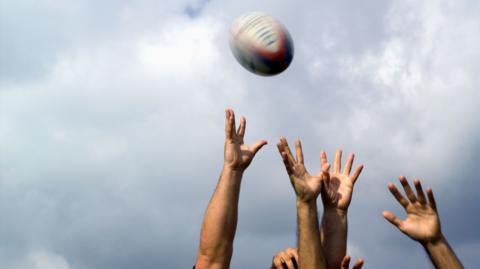 Hands rreaching for a rugby ball which is up in the air. The background is the sky with clouds.