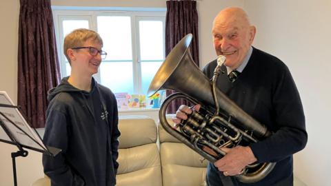 Neythen is standing in a living room next to Brian Routledge, who is holding a euphonium. They are both smiling.