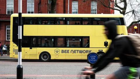 A cyclist passes a yellow-liveried Bee Network, double decker bus across a street in the day time in Manchester.