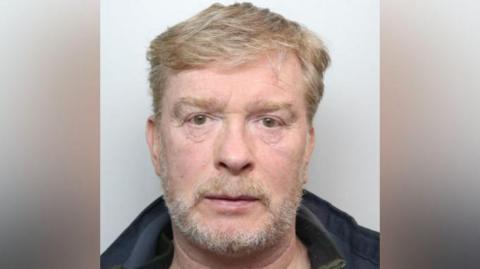 Mugshot of Aron Traynor who has short sandy-coloured hair and beard. He wears a dark jacket