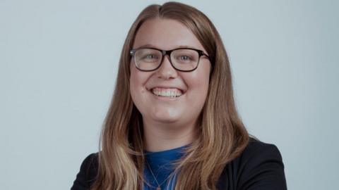 Rachel Hayden smiles at the camera. She has long dark blonde hair, light blue eyes, and wears brown-rimmed glasses.