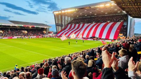 Pittodrie Stadium - Aberdeen FC's Pittodrie Stadium, full with thousands of fans, enormous red and white banner covering almost all of one stand, hands in air clapping, green pitch and blue early evening sky and floodlights.