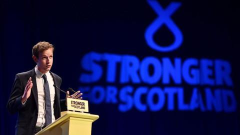 Callum McCaig wearing a dark suit over a white shirt and dark tie. He is standing behind a podium. The words 'Strong for Scotland' are in large blue letters behind him.