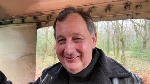 A man wearing a black jacket stands and smiles at the camera. Woodland can be seen behind him.
