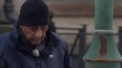 An elderly man using a walking stick and wearing a black hat and long coat walking towards court with help from someone who is blurred out.