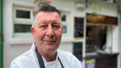 A man with grey hair, wearing a white chef shirt and a dark blue apron, is looking into the camera. He's standing in front of the cafe he owns, and there are two black boards on the white building wall behind him and a hatch for serving.